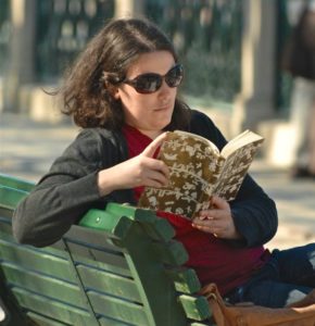 Dark haired woman wearing sunglasses reading a book, sitting on a park bench