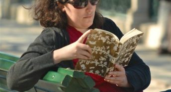 Dark haired woman wearing sunglasses reading a book, sitting on a park bench