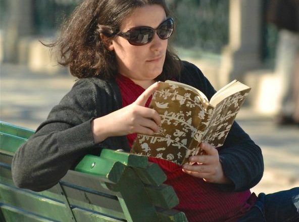 Dark haired woman wearing sunglasses reading a book, sitting on a park bench